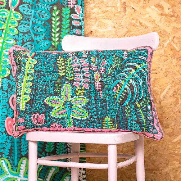 Decorative needlepoint cushion with botanical pattern on a pink chair against a colourful fabric and wood background