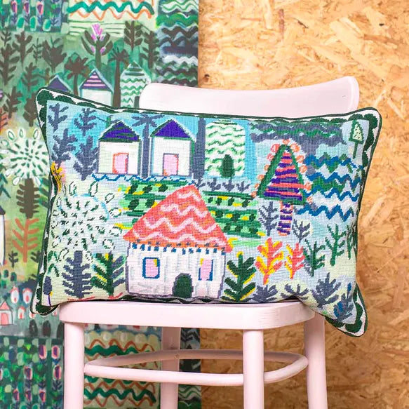 Decorative needlepoint cushion with colourful house design on a chair against a wooden wall