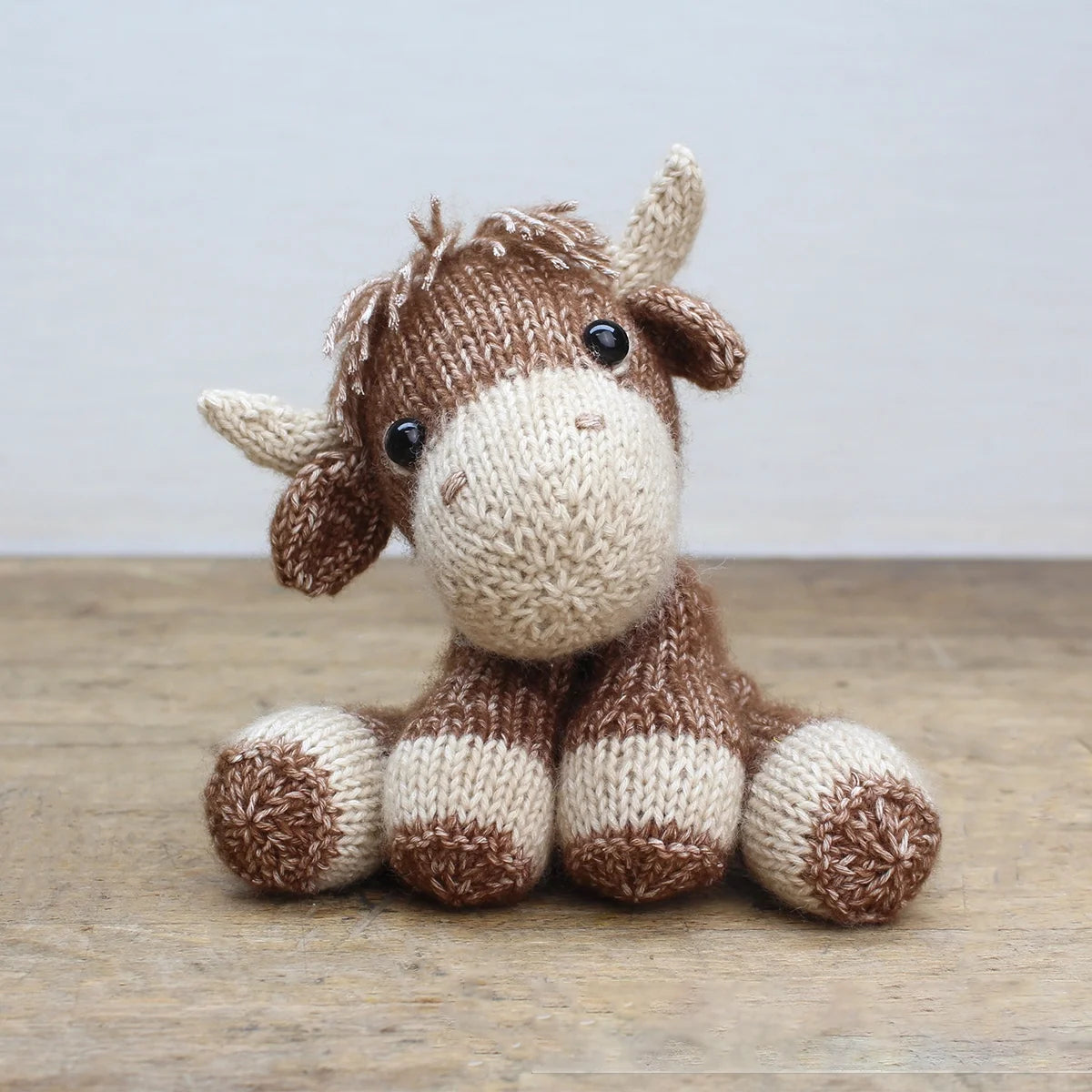Highland cow soft toy knitted in cotton-blend yarn on a wooden surface with light grey background