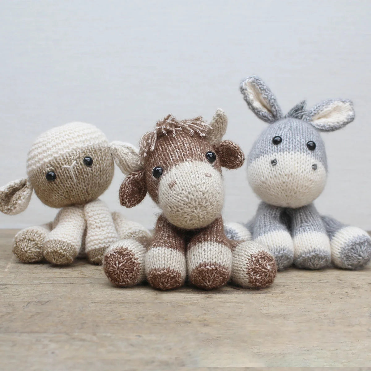 Three farmyard knitted animal soft toys on a wooden surface with a light grey background