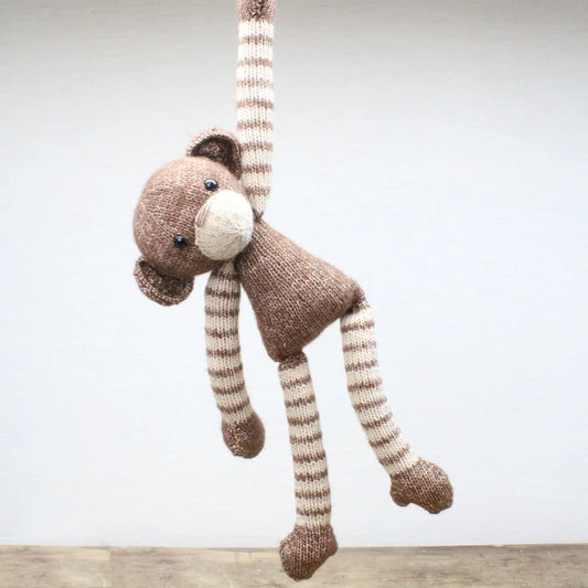 Knitted brown monkey toy with striped legs hanging against a plain background