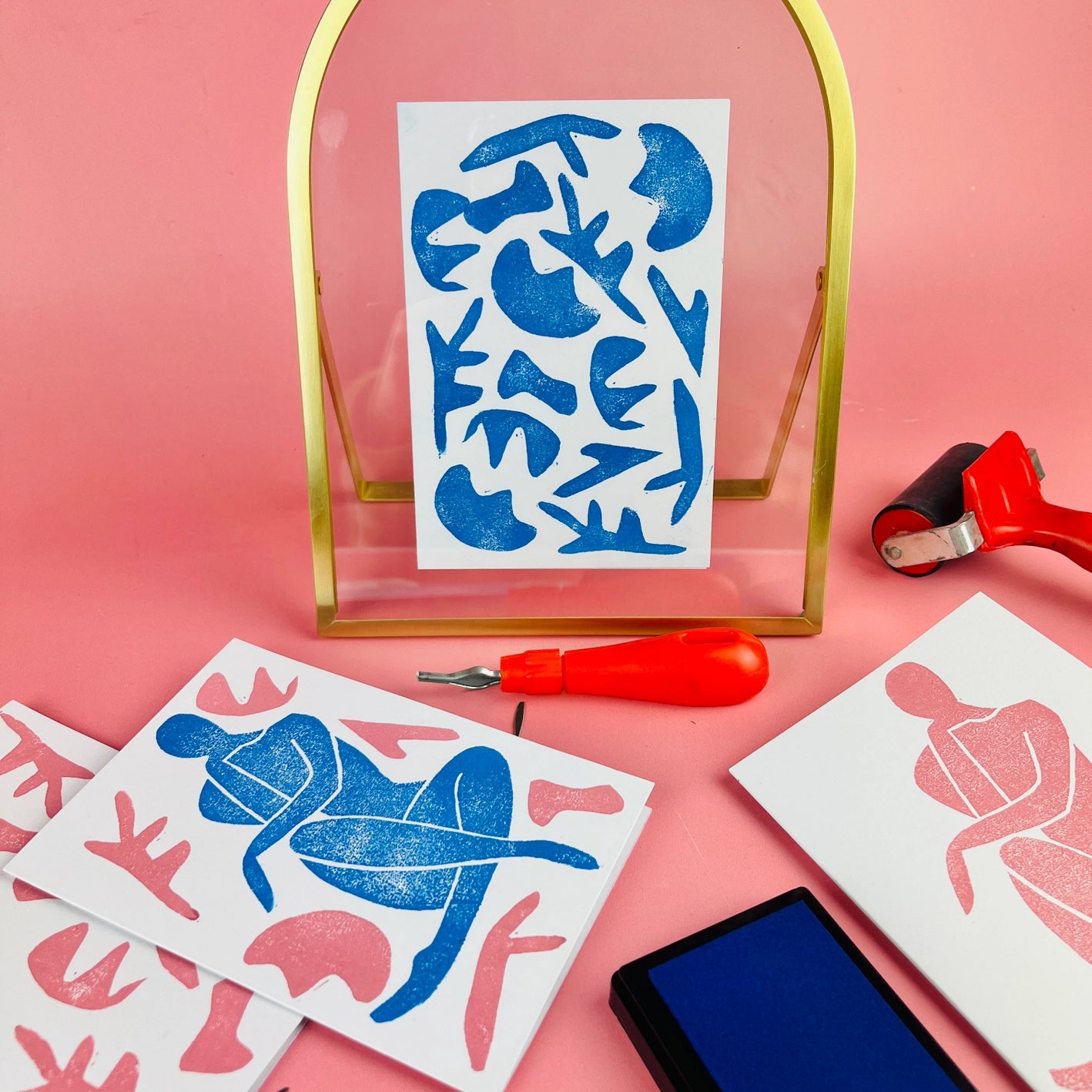 A collection of craft materials for lino printing arranged on a pink surface, including a cutter, a blue ink pad, and printed cards with Matisse-inspired designs
