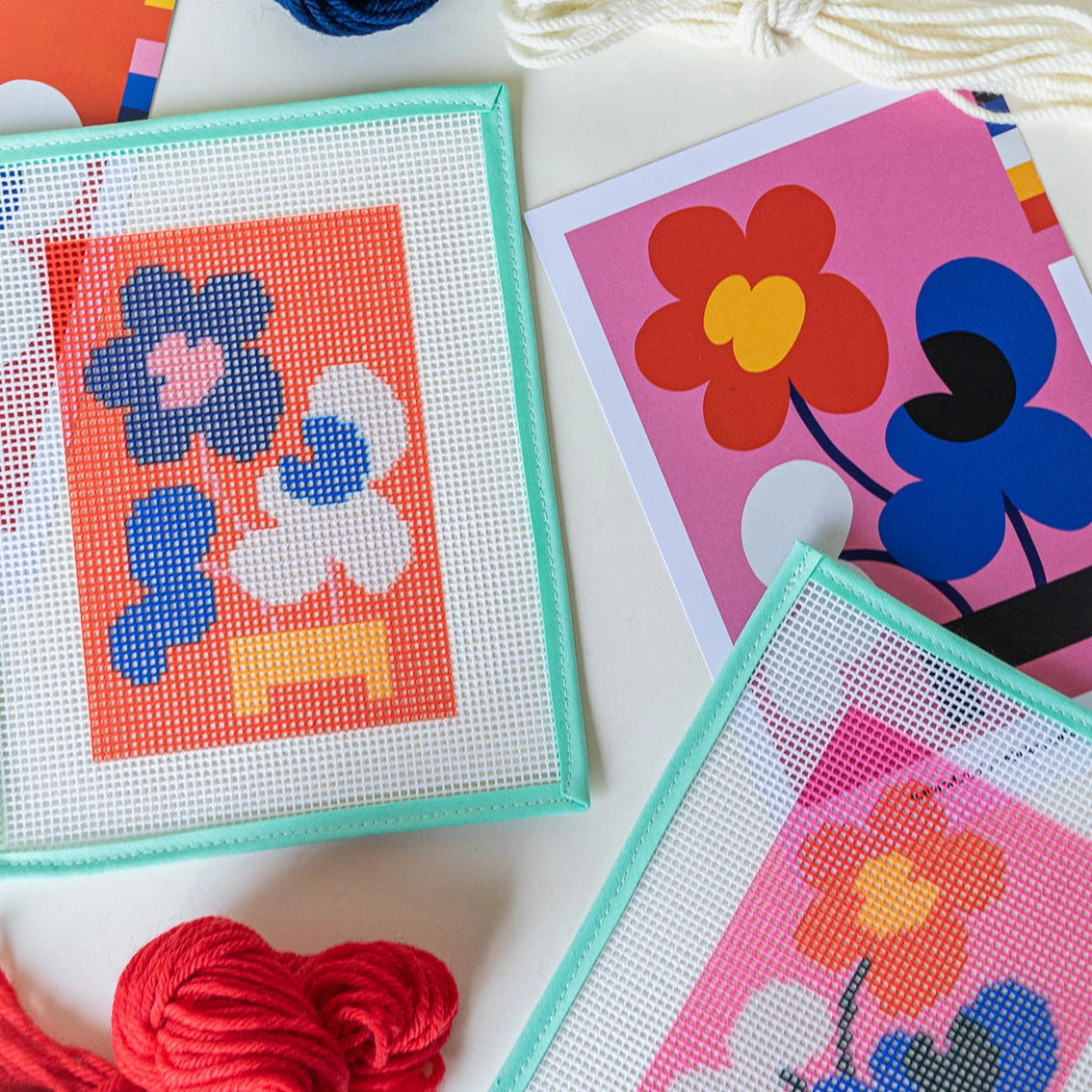 Two printed needlepoint canvases, each featuring a simple floral design inspired by ikebana, alongside the original artwork and skeins of coloured yarn