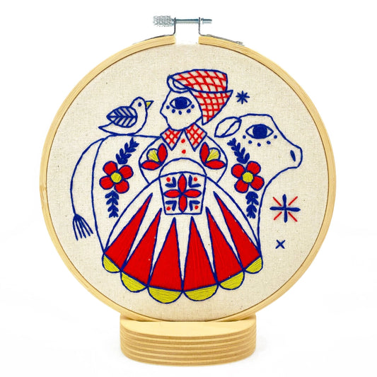 Folk-style embroidery of a milkmaid in a red, blue and yellow dress with a blue cow and bird, surrounded by floral motifs
