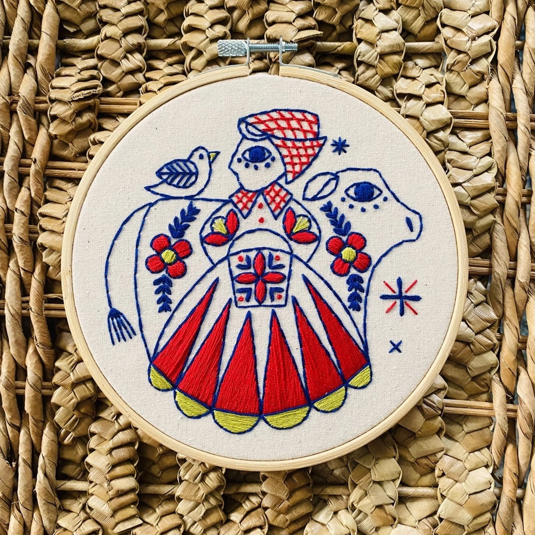 Folk-style embroidery of a milkmaid in a red, blue and yellow dress with a blue cow and bird, surrounded by floral motifs on a straw background