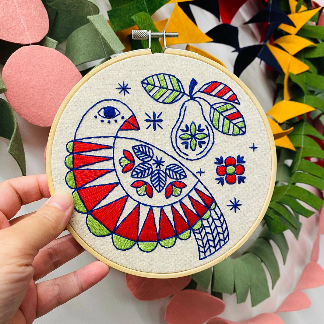 Hand holding a completed partridge and pear embroidery in a colourful Scandi folk style, set against a bright background