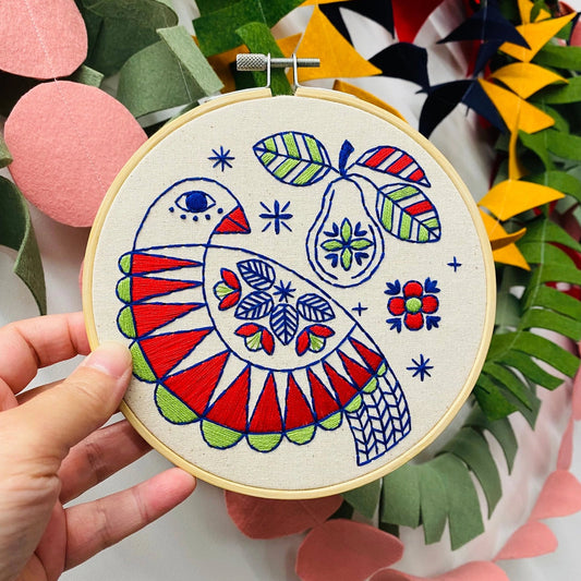 Hand holding a completed partridge and pear embroidery in a colourful Scandi folk style, set against a bright background