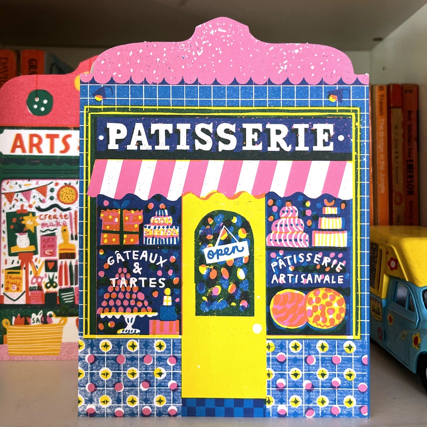 A colourful art card featuring a patisserie shop front with a vibrant window display, set against a background of another shop front card and toy ice cream van