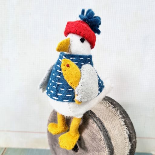 Felt seagull wearing a red hat and blue clothing, holding a yellow fish
