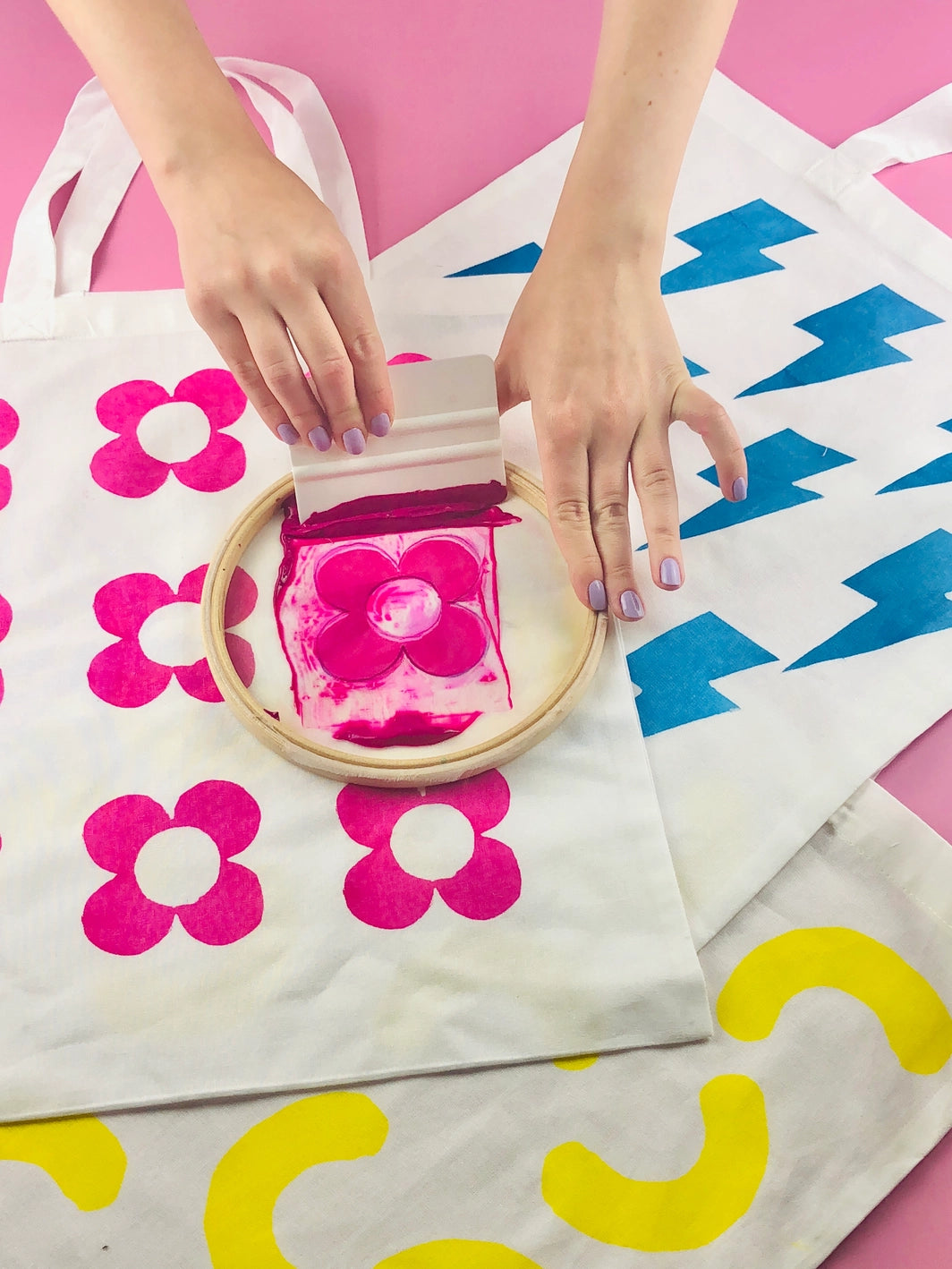 A person screen printing pink floral designs on a white tote bag using a bamboo embroidery hoop and a squeegee, on a background of other screen printed tote bags