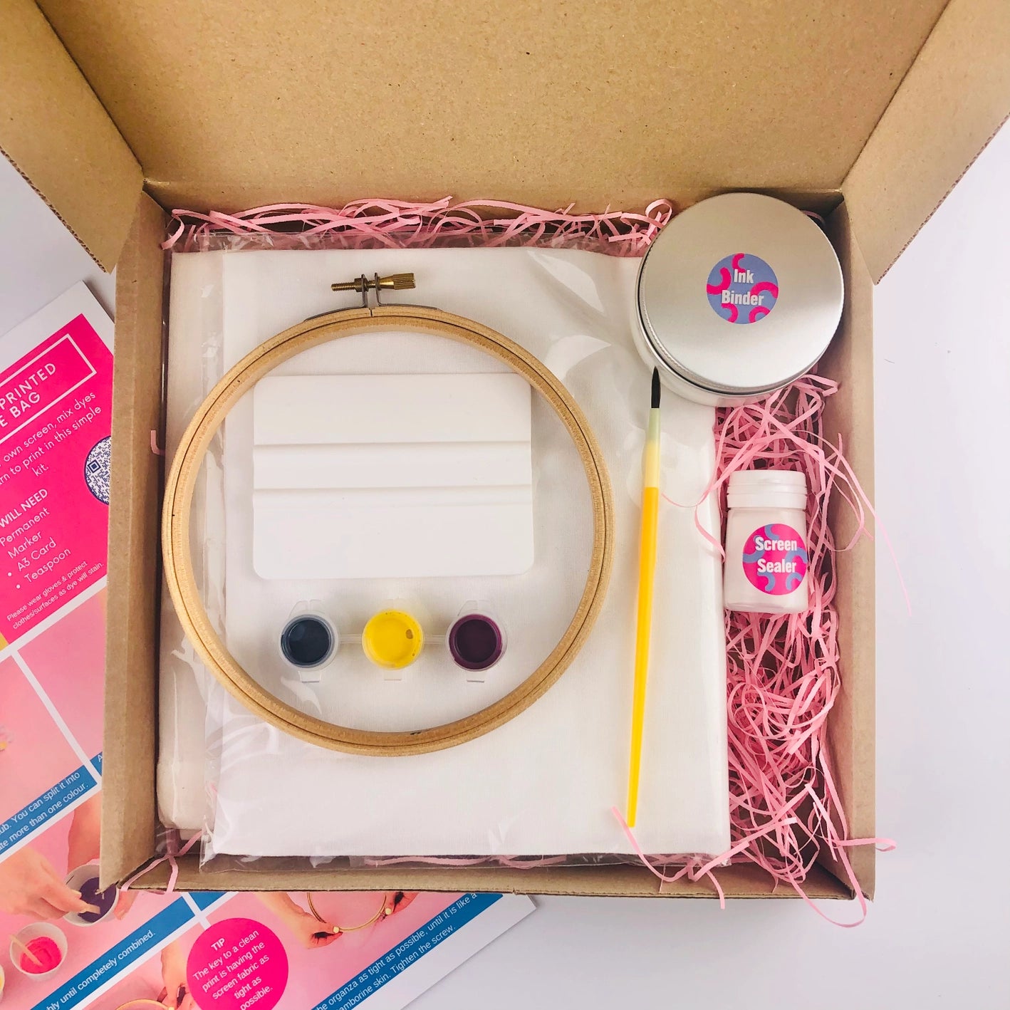 Close-up of screen print tote bag kit box contents, including tote bag, embroidery hoop, inks, ink binder, screen sealer and paint brush, with instruction sheet on a pink paper straw background