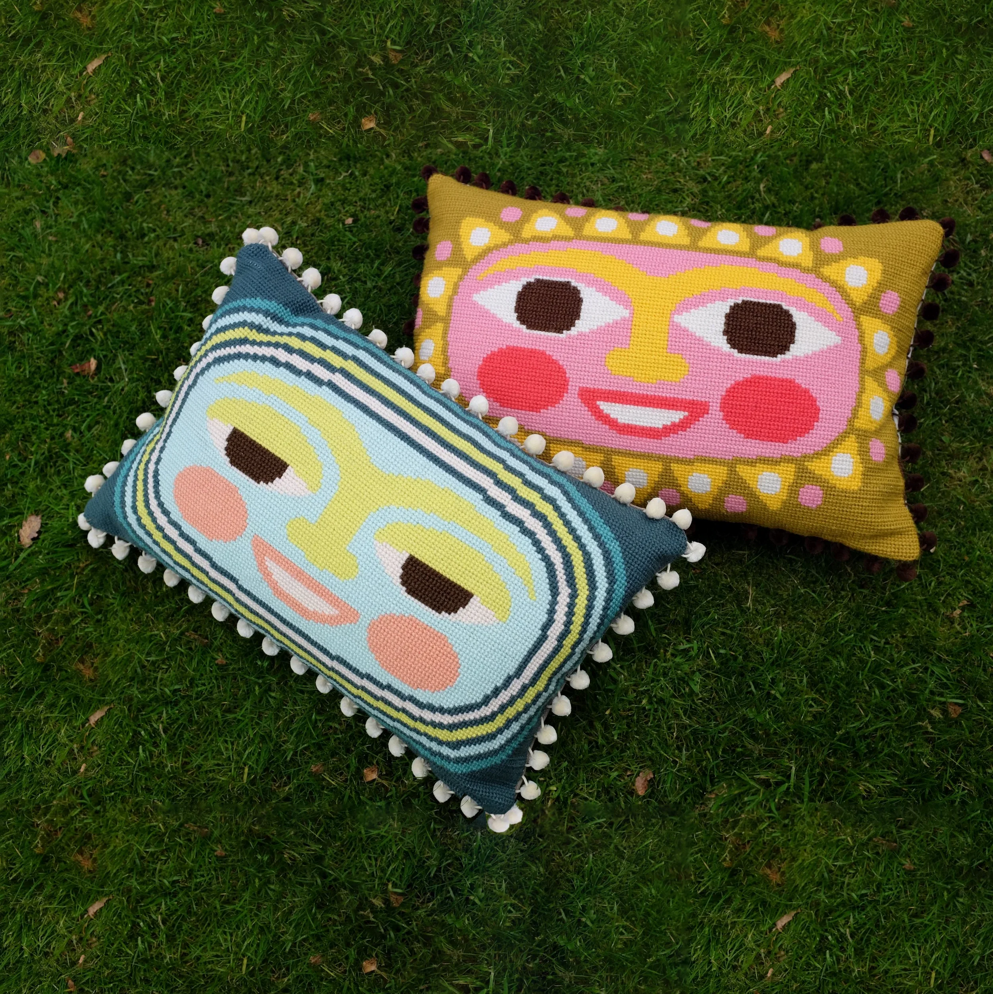 Two decorative needlepoint pillows with solar and lunar face designs on a grassy background