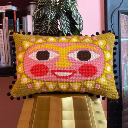 Needlepoint sun face pillow in warm pink and yellow tones on a gold stool with plant and books in the background