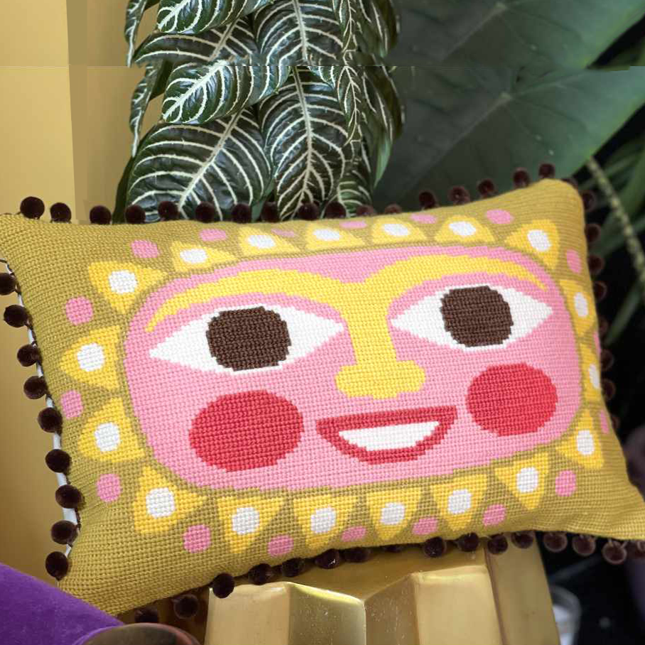 Decorative needlepoint sun face pillow in warm pink and yellow tones on a gold stool with plant and books in the background