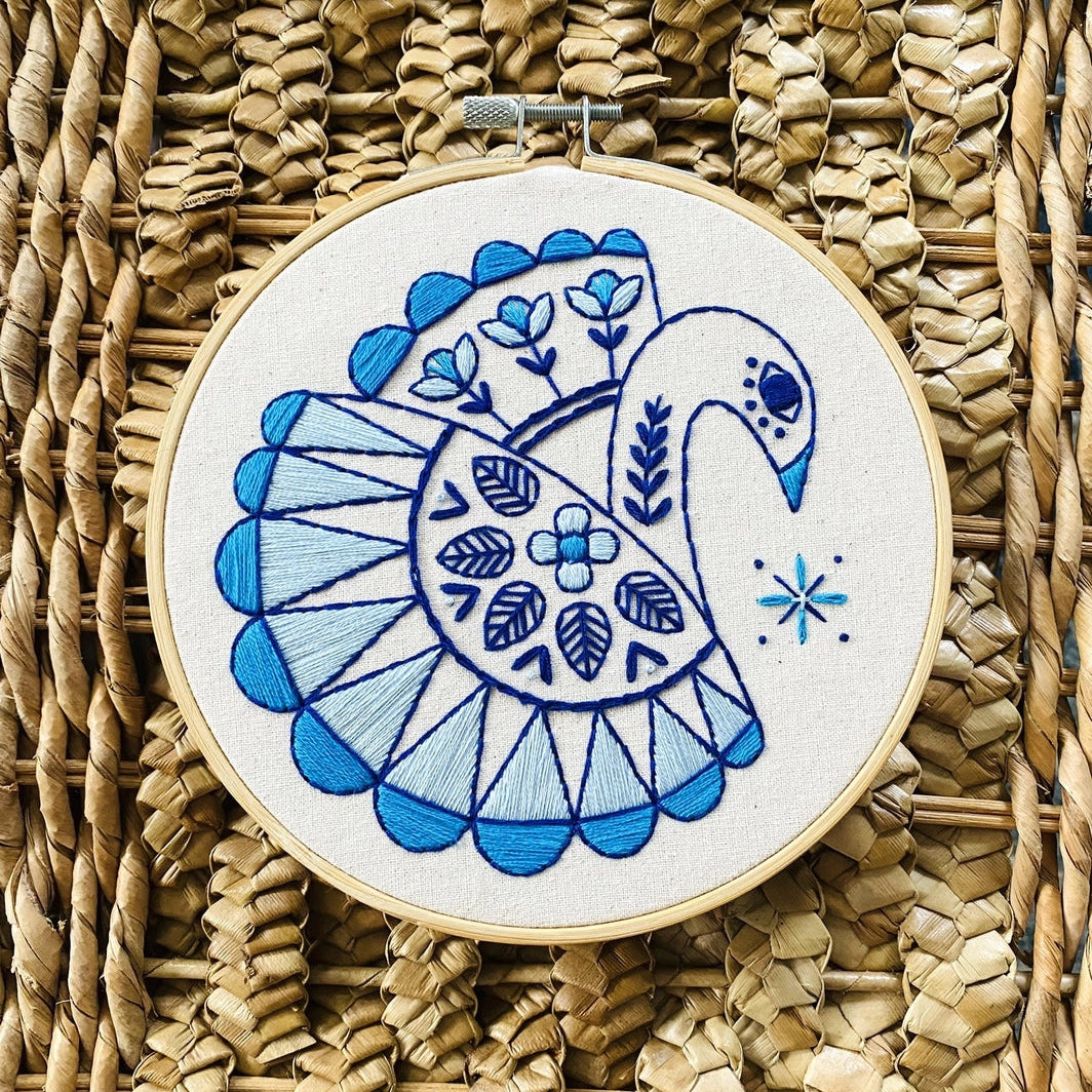 Embroidery hoop with a blue and white swan design on unbleached cotton, displayed on a woven mat