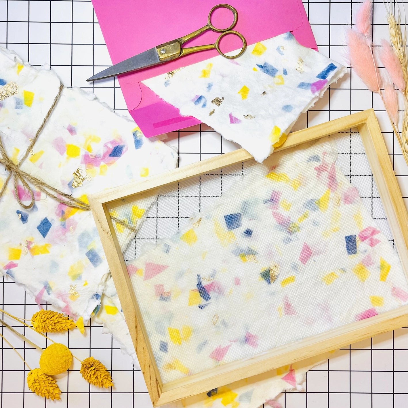 Handmade pieces of terrazzo paper and paper making kit tools, including a mesh frame and pair of scissors on a grid background