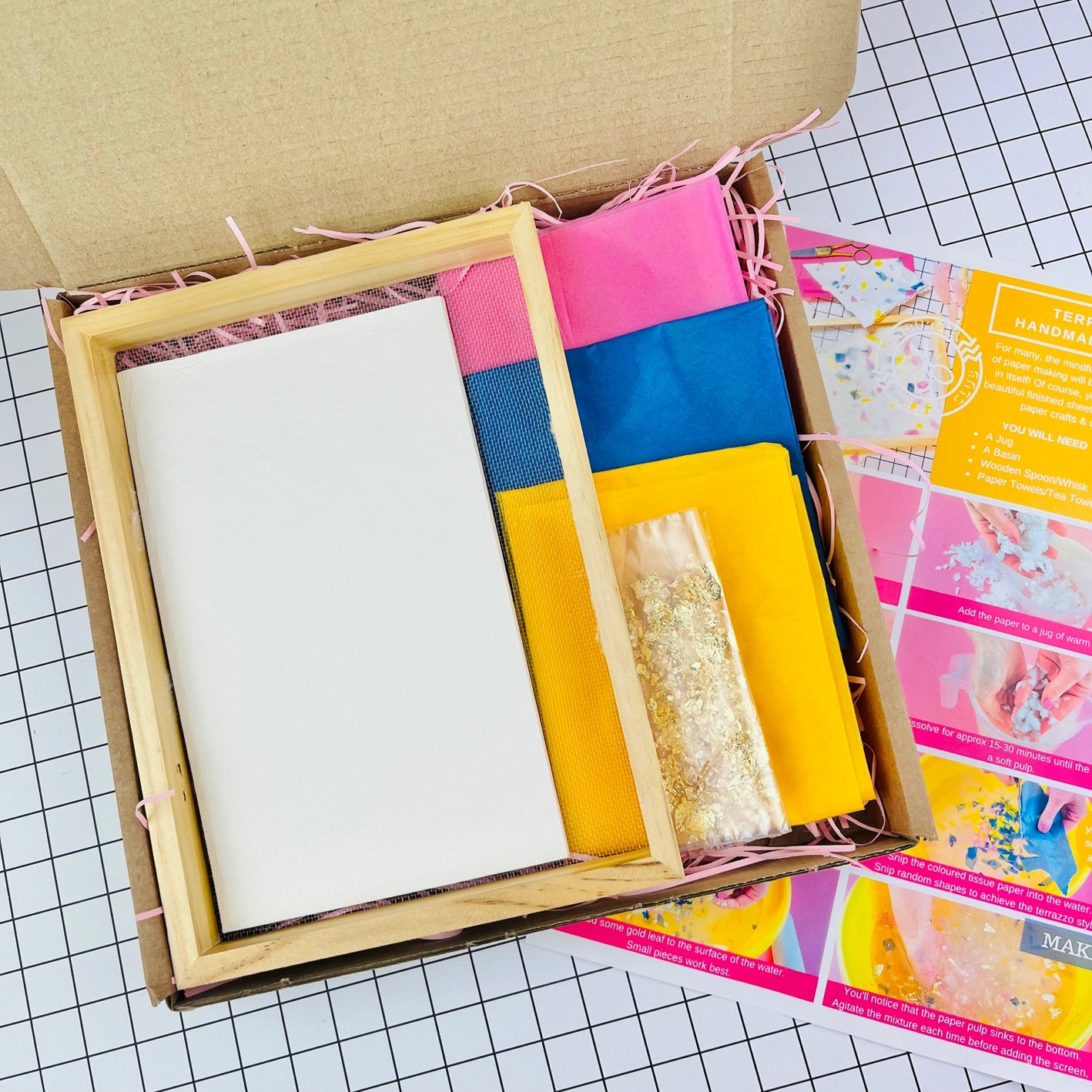 Close-up of terrazzo paper making kit box contents, including brightly coloured tissue paper, mesh frame, plain paper and gold leaf, with instruction sheet on a grid background