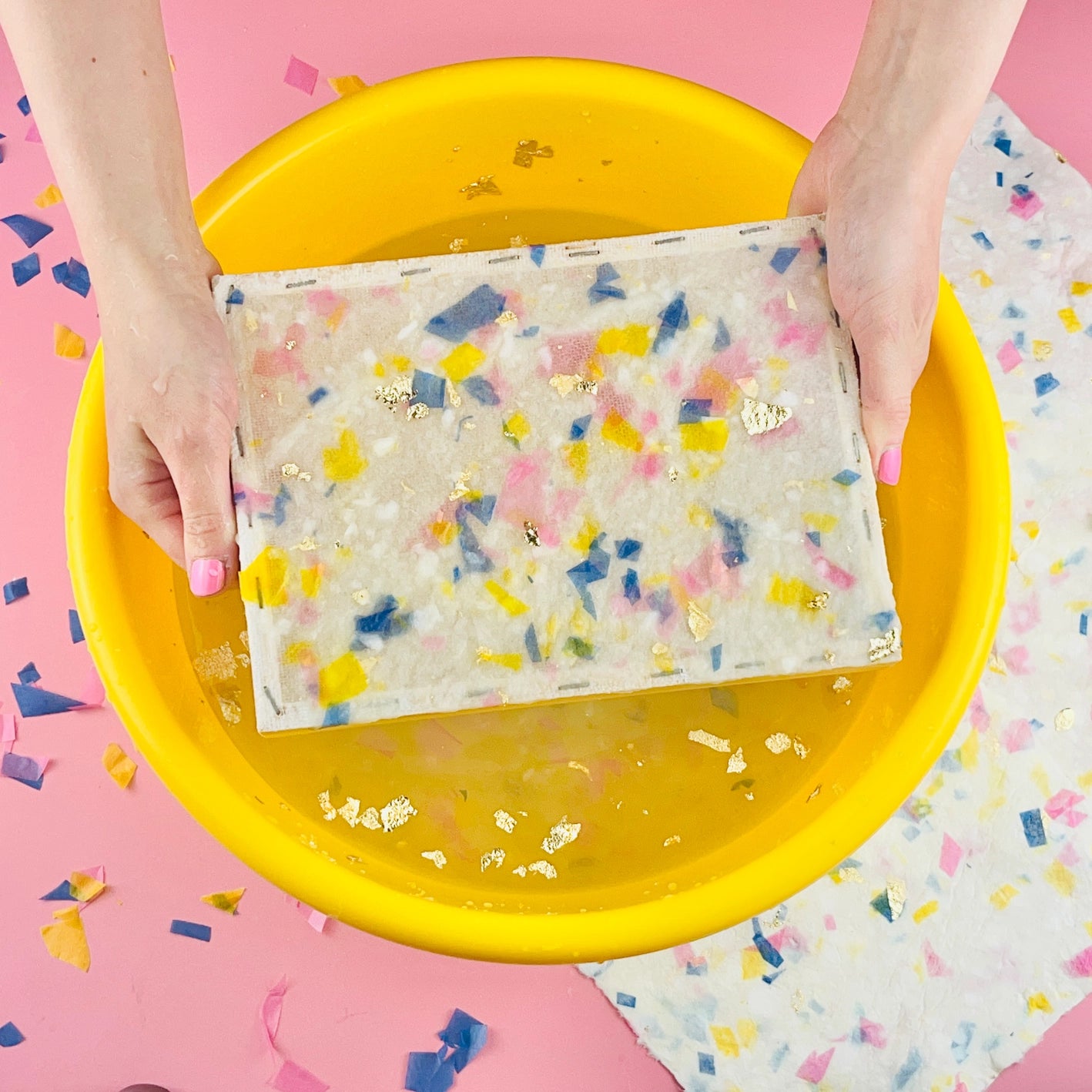 Close-up of person creating terrazzo paper using a mesh frame in a plastic yellow bowl on a pink background