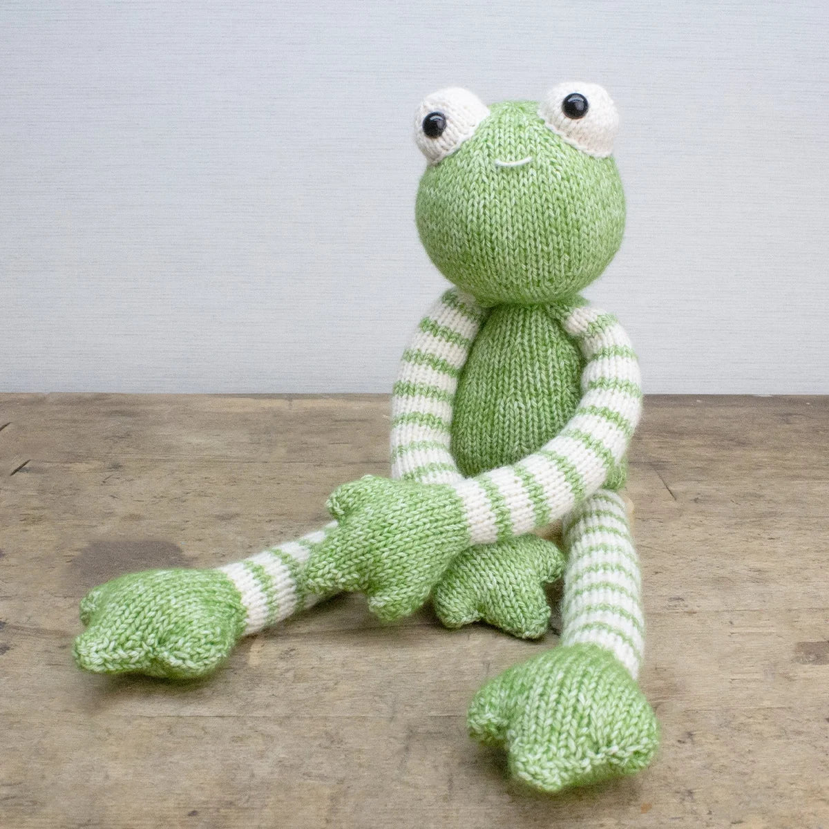 Knitted green and white frog soft toy on a wooden surface with a light grey background
