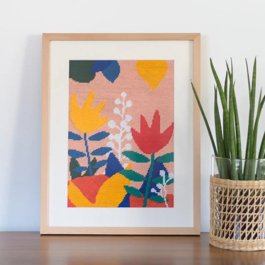 Framed needlepoint depicting colourful floral patterns displayed on a table alongside a plant